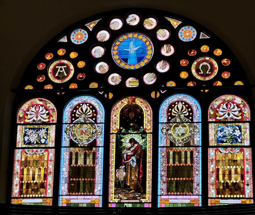 Photo of a stained glass window in a church. The space is dark and the light shining from the outside illuminates the designs of the stained glass window.