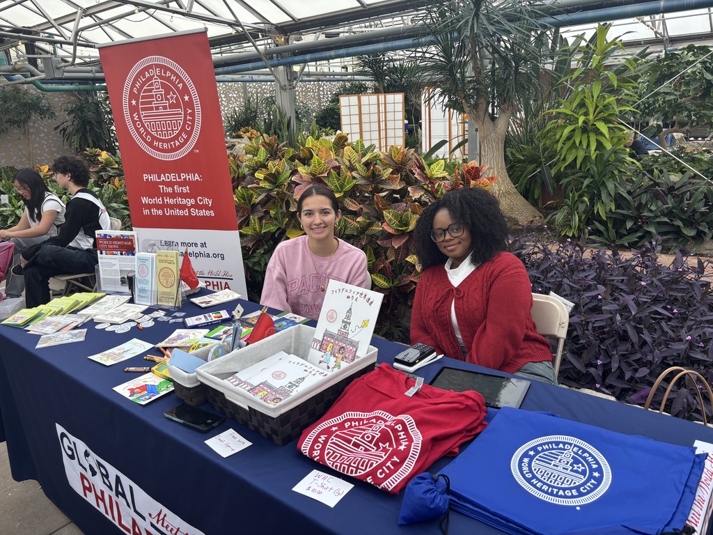 Two women are seated at a table smiling at the camera. On the table there are various items such as coloring books, shirts, totes, pamphlets and signs all with the World Heritage City Seal on them. Behind them is a poster standing up with the World Heritage Seal displayed on it.